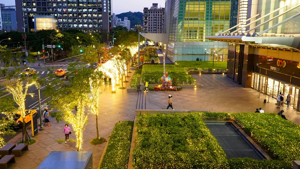 Commercial plaza at dusk with illuminated trees, landscaped beds and wide pedestrian paths